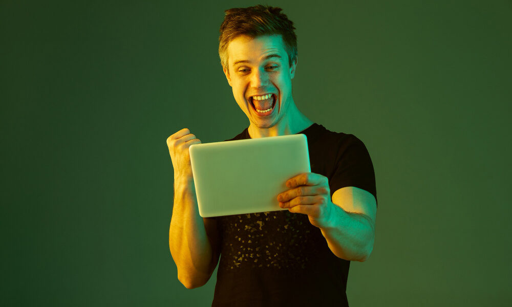 holding tablet celebrating win bet game caucasian man s portrait green studio background neon light beautiful male model concept human emotions facial expression sales ad